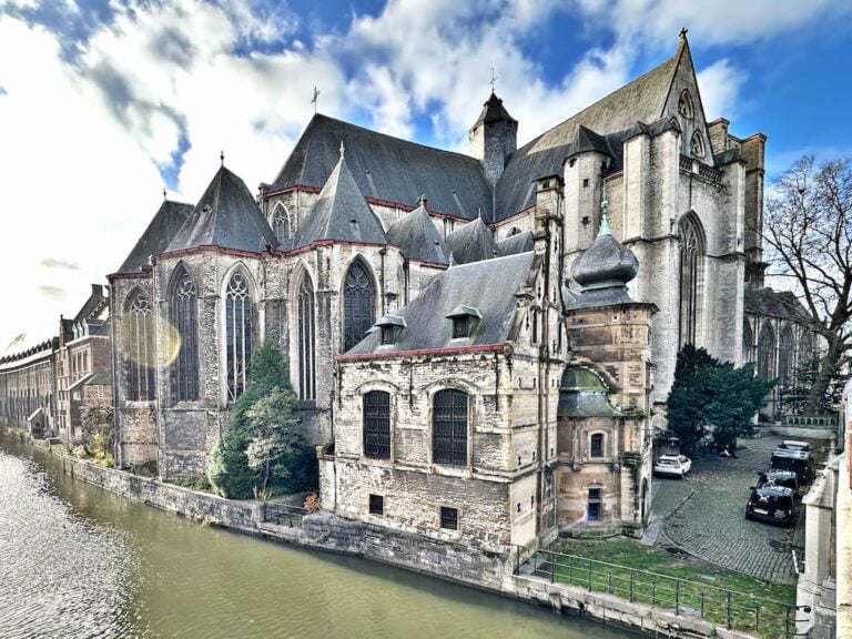 Large Gothic-style stone church with pointed arches and buttresses, located beside a canal on a sunny day with scattered clouds. Cars are parked near the building.