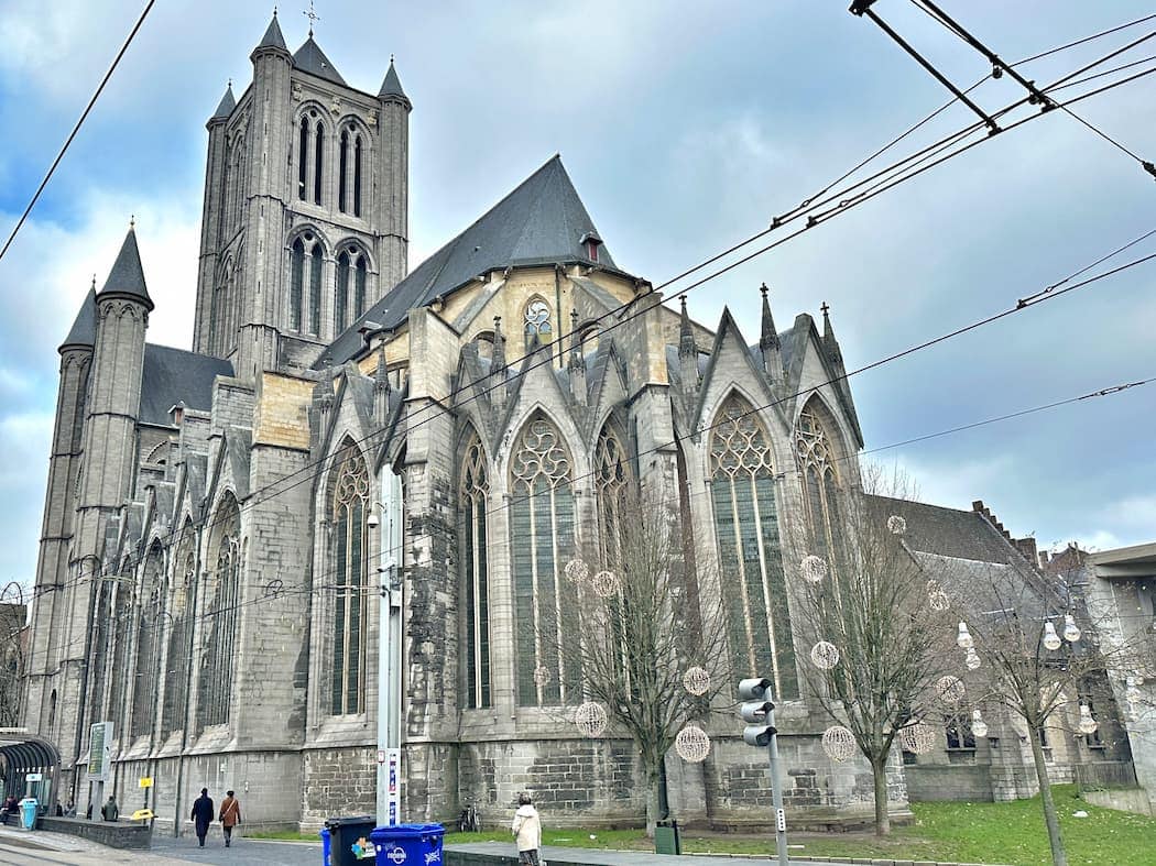 A large stone Gothic church with pointed arches and tall stained glass windows, seen from the street with a few pedestrians and tram wires overhead.