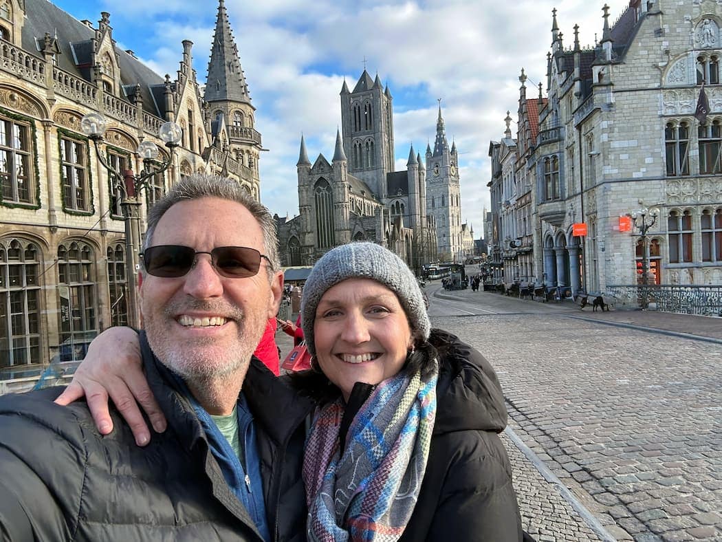 A man and woman smile for a selfie on a cobblestone street lined with historic buildings and a large Gothic church in the background.