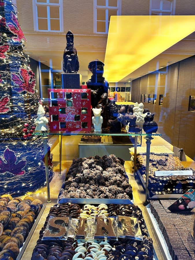 Assorted chocolates, chocolate figurines, and a chocolate letter spelling "SINT" are displayed in a shop window with festive decorations and reflections.