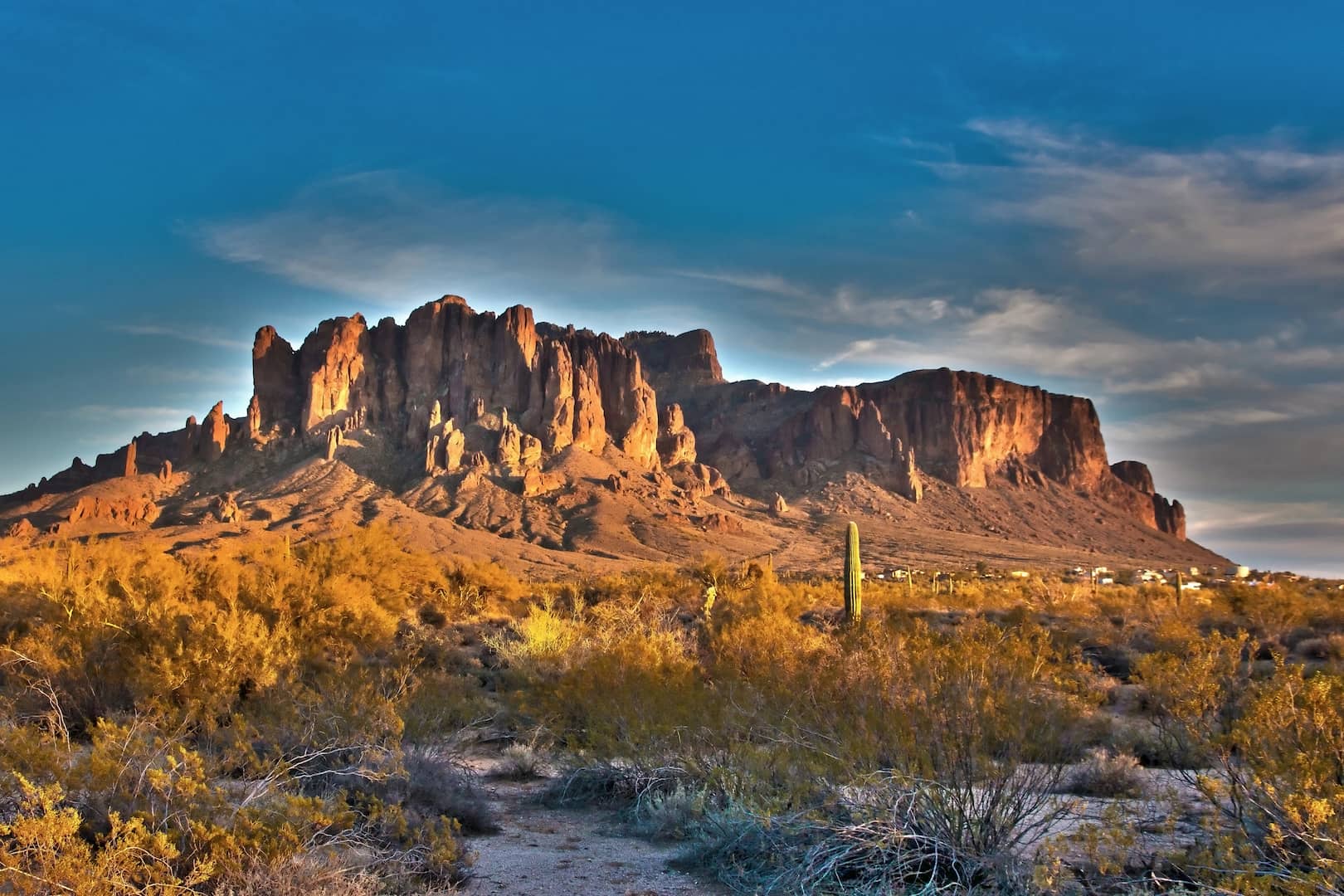 A rocky mountain range rises from a desert landscape under a blue sky with scattered clouds; sparse vegetation and a few cacti are visible in the foreground—perfect scenery for bucket list trips or relaxing vacation homes.
