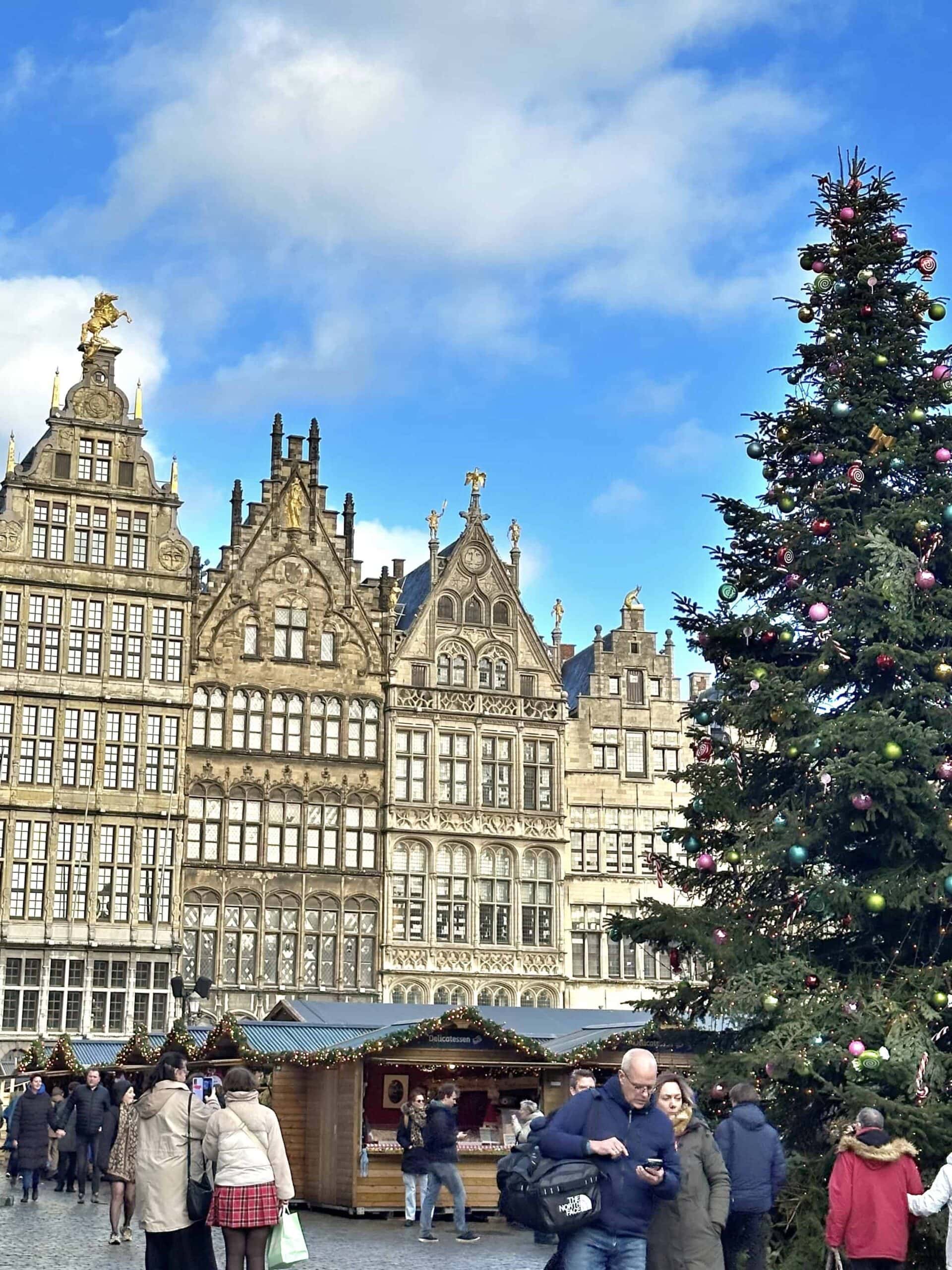 People walk in a town square decorated for Christmas, with historic buildings in the background and a large decorated Christmas tree on the right.