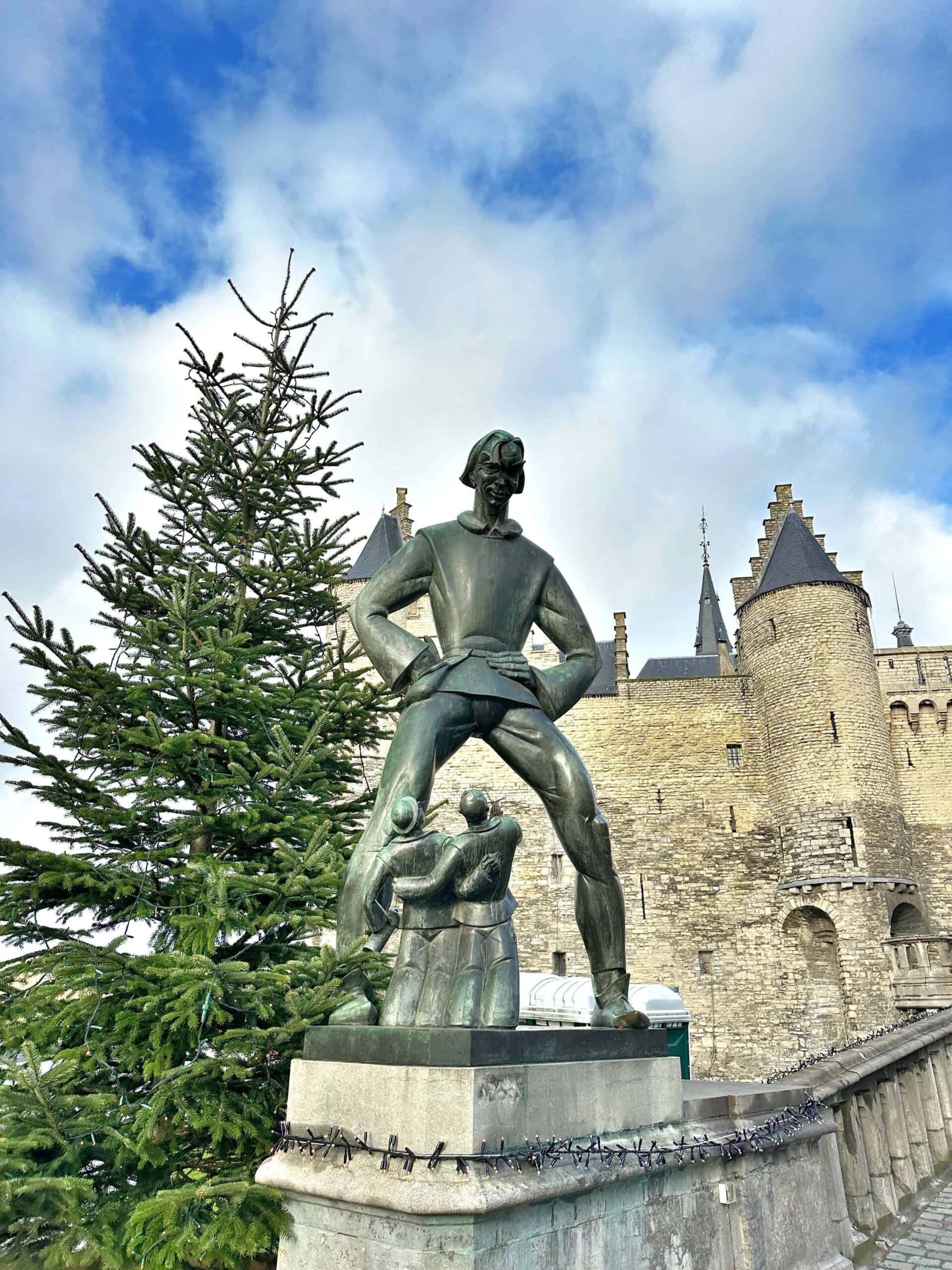 Bronze statue of a man with hands on hips beside two smaller figures, next to a pine tree, with a stone castle and cloudy sky in the background.