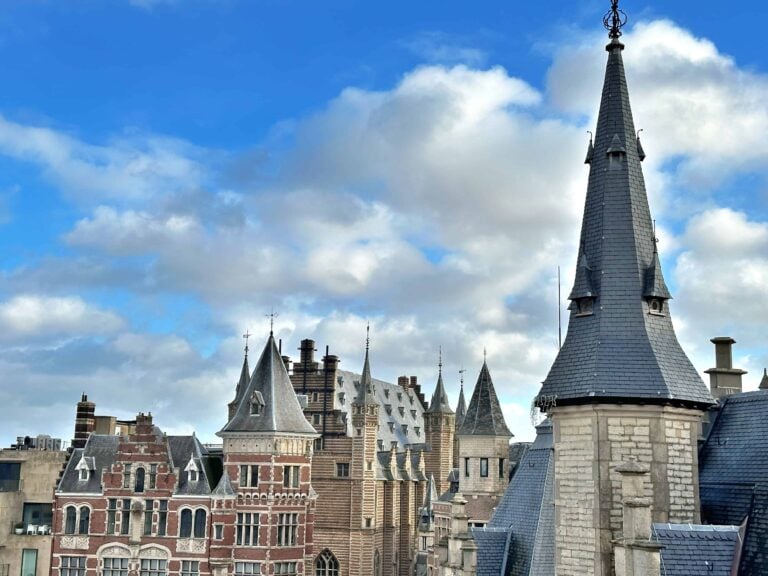 Several ornate, historic buildings with pointed spires and intricate brickwork stand under a partly cloudy blue sky.
