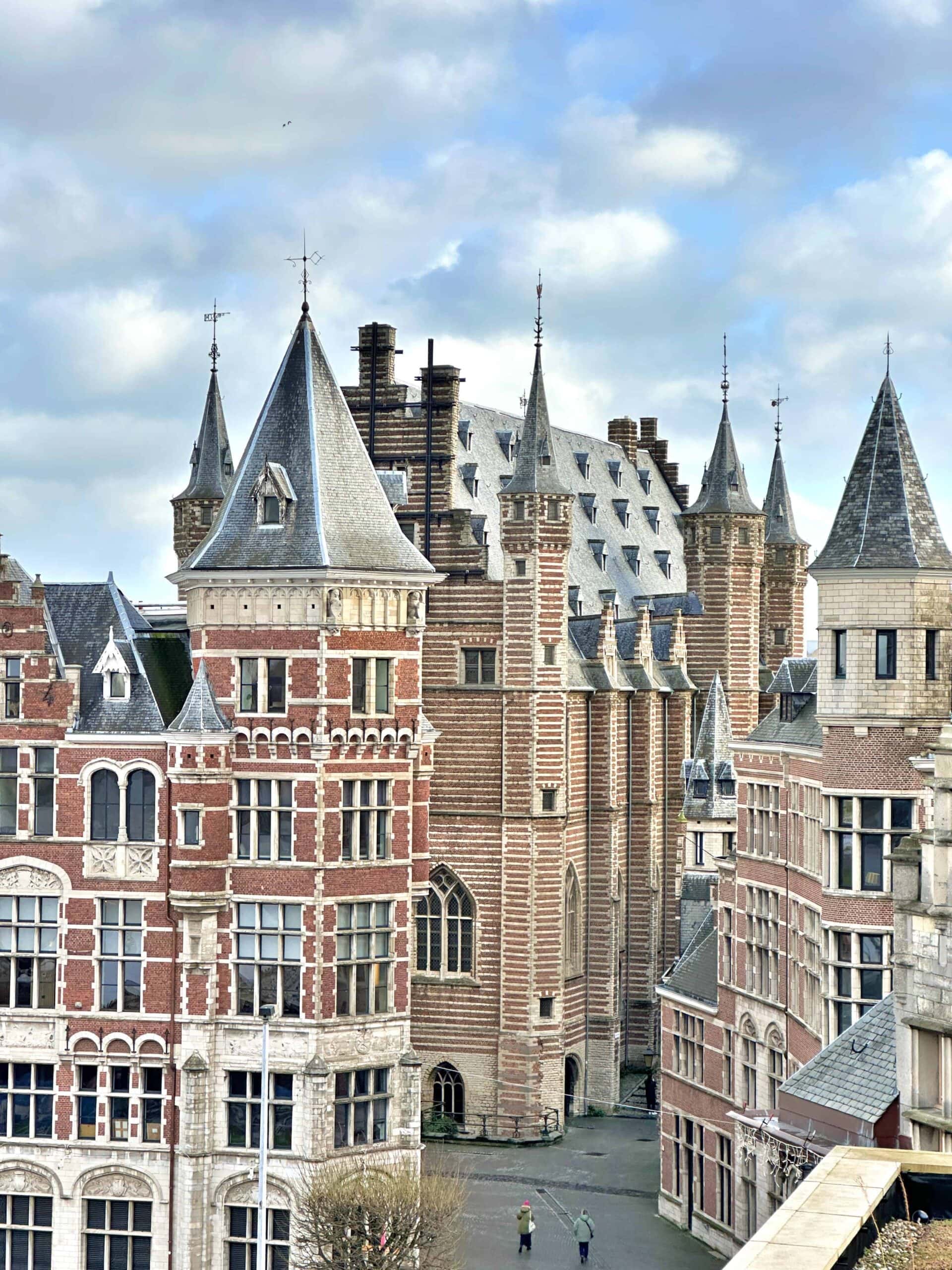 View of historic European buildings with ornate brickwork, pointed rooftops, and chimneys under a partly cloudy sky, with two people walking on a street below.