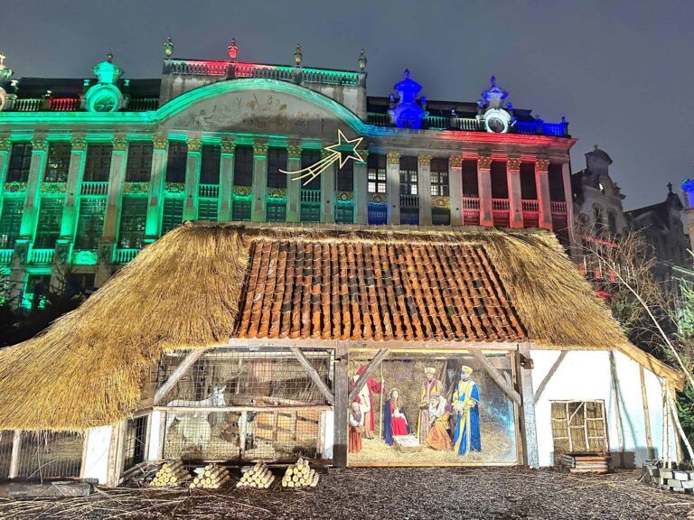 A nativity scene with figures is displayed in front of a thatched-roof structure; a large building behind is lit with green, red, and white lights.