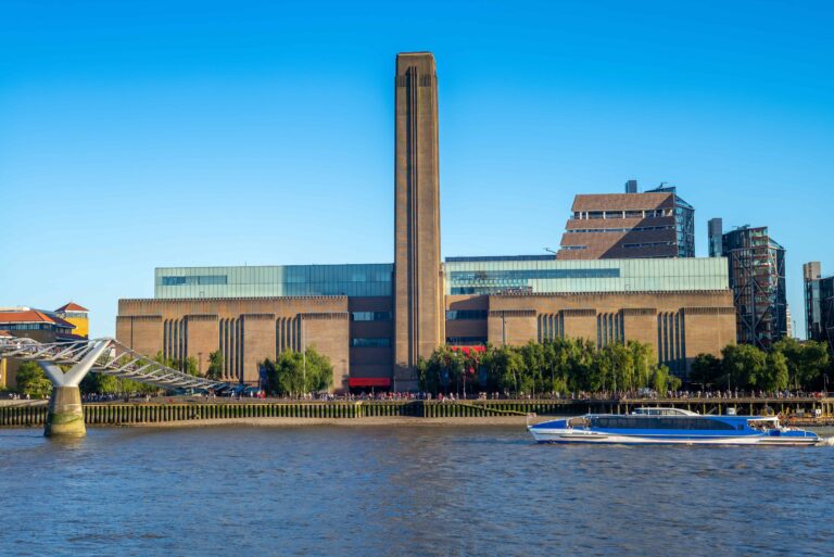 The image shows the Tate Modern art museum, one of the top London Museums that are Free, along the River Thames, with the Millennium Bridge to the left and a boat on the river in the foreground.