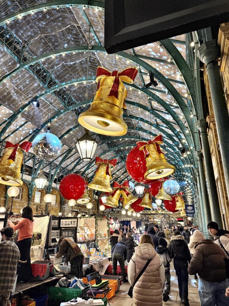 Indoor market decorated with large gold bells, red ornaments, and lights captures the festive spirit of Christmas Markets in London. Shoppers browse various stalls under a glass roof adorned with holiday decorations.