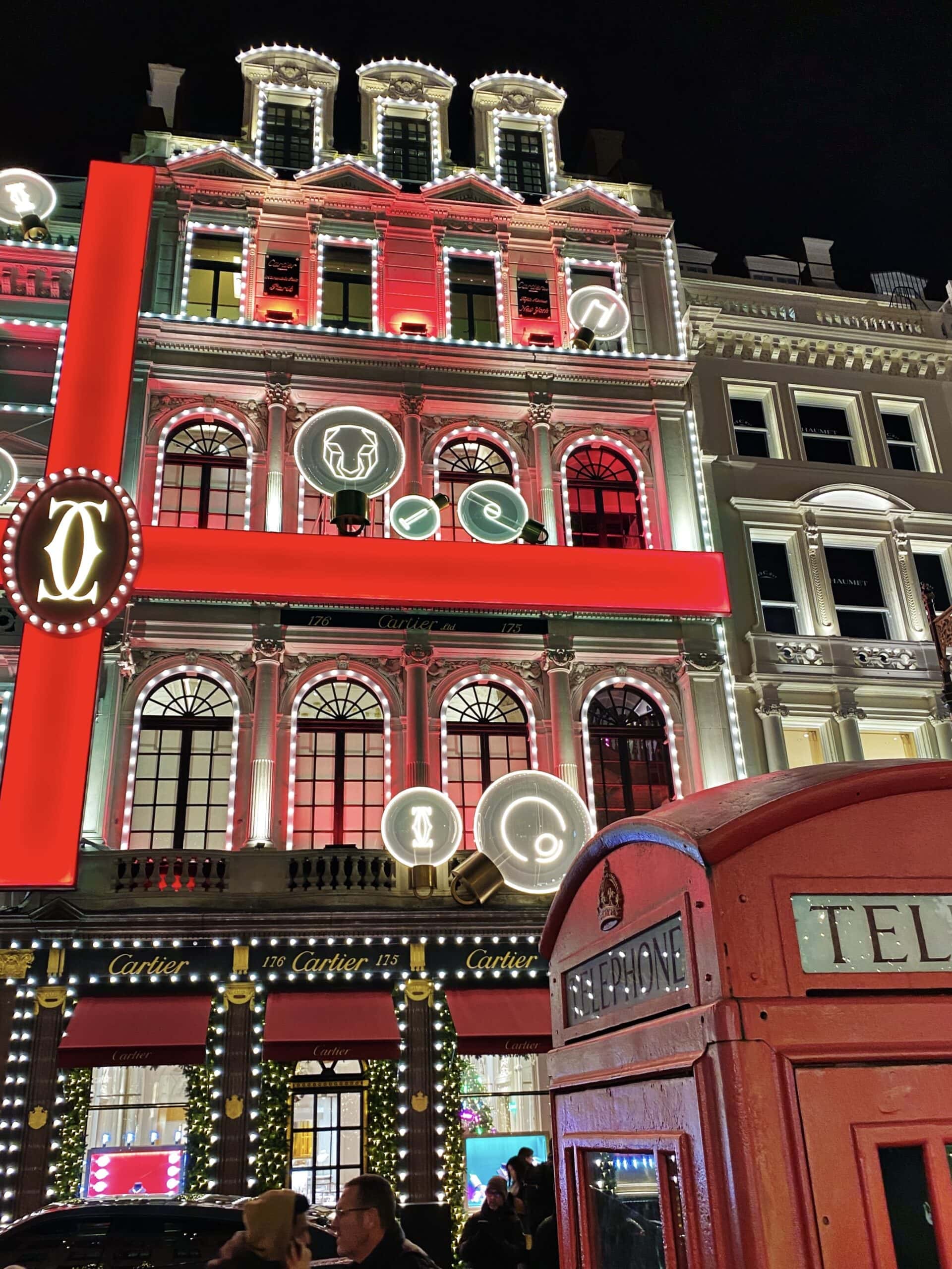 A brightly lit Cartier store decorated with red lights and symbols at night, alongside a classic red British telephone booth in the foreground, captures the festive spirit of Christmas lights in London.