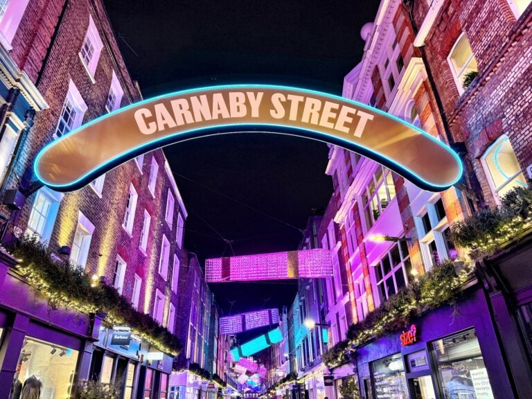 A brightly lit Carnaby Street sign arches over a lively, colorful street at night, where Christmas Lights in London twinkle alongside illuminated decorations and historic brick buildings on both sides.