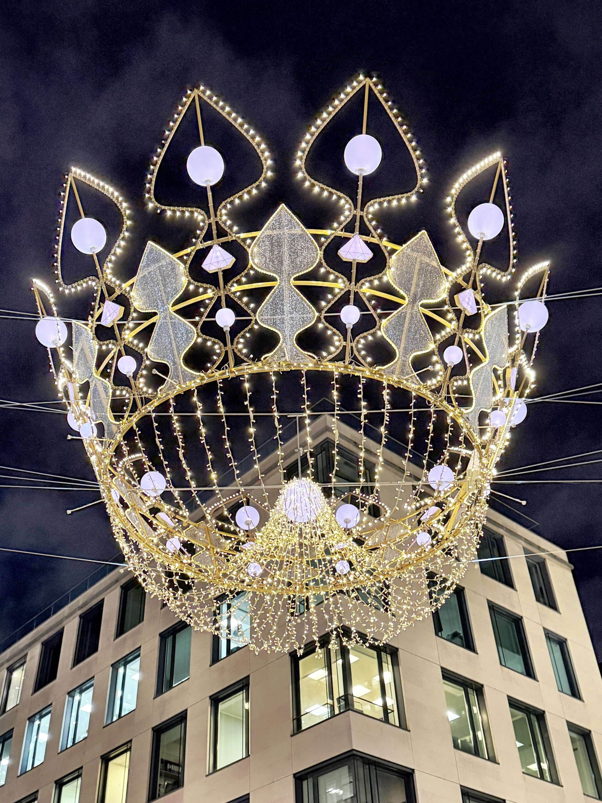A large illuminated crown-shaped decoration, part of the Christmas Lights in London, is suspended above a city street, with a modern building and night sky in the background.