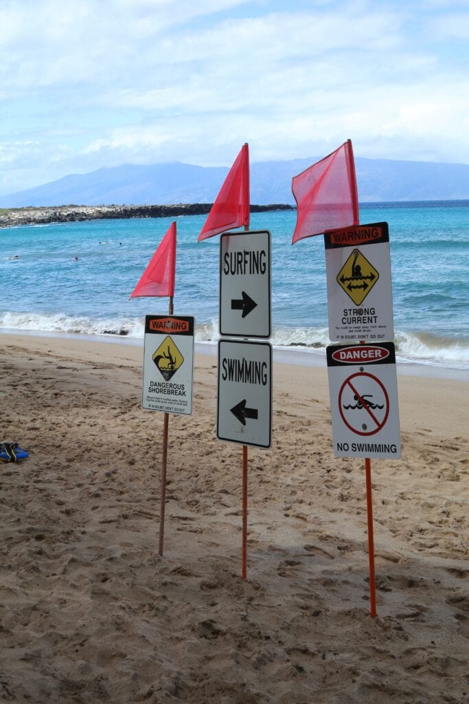 Warning signs with red flags at DT Fleming Beach indicate dangerous shorebreak, strong currents, and no swimming areas; surfing, swimming, and DT Fleming Beach snorkeling directions are also posted.