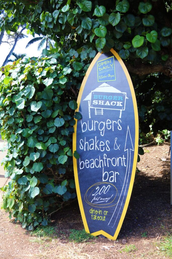 A surfboard-shaped sign advertises "Burger Shack," serving burgers, shakes, and a beachfront bar near DT Fleming Beach Snorkeling, open daily 11am-3pm for dine-in or takeout.