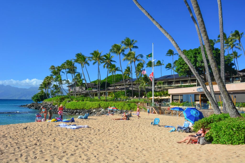 People relax on the sandy beach at Napili Bay, with palm trees, umbrellas, and resort buildings in the background—perfect for a sunny day or snorkeling adventures in Maui under a clear blue sky.