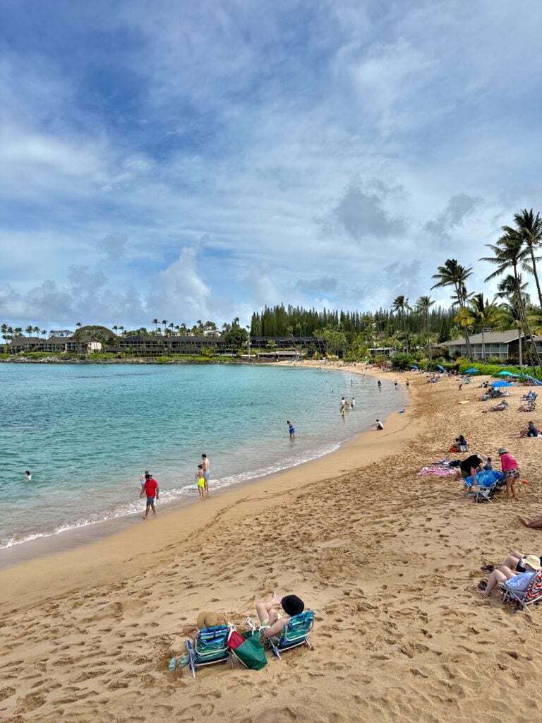 People relax on the sandy beach at Napili Bay, with clear blue water perfect for snorkeling, palm trees, and resort buildings in the background under a partly cloudy Maui sky.