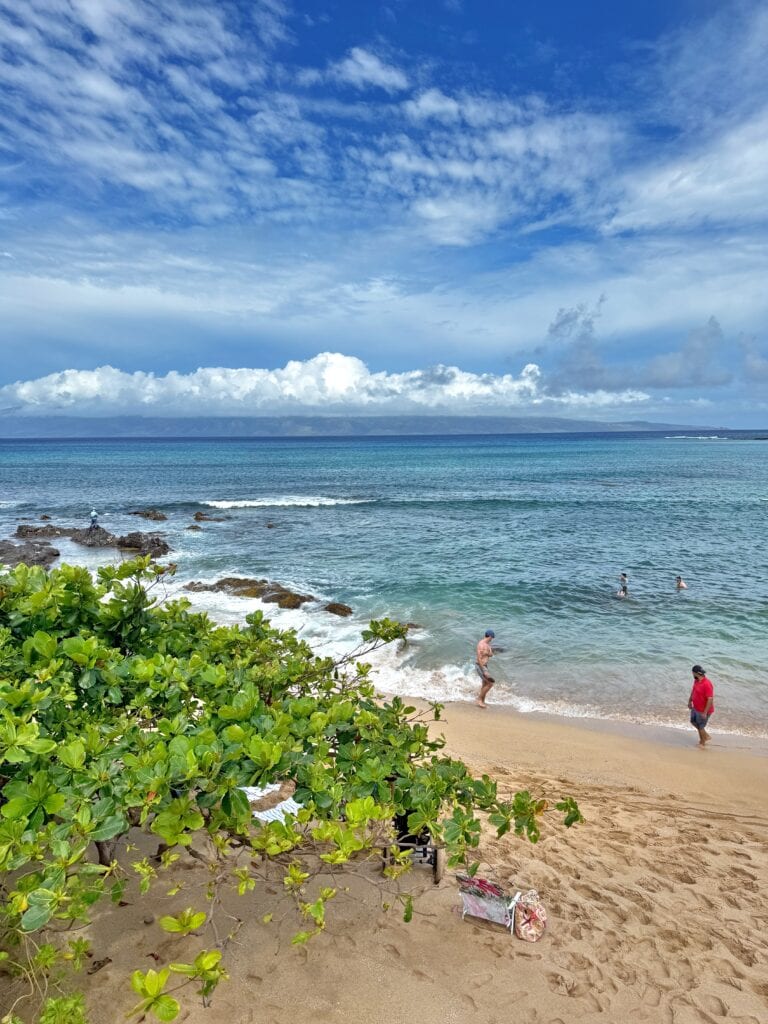 People are walking and swimming at the sandy shores of Napili Bay, Maui, enjoying clear blue water and partly cloudy skies. Green foliage frames the view, perfect for snorkeling or relaxing by the beach.