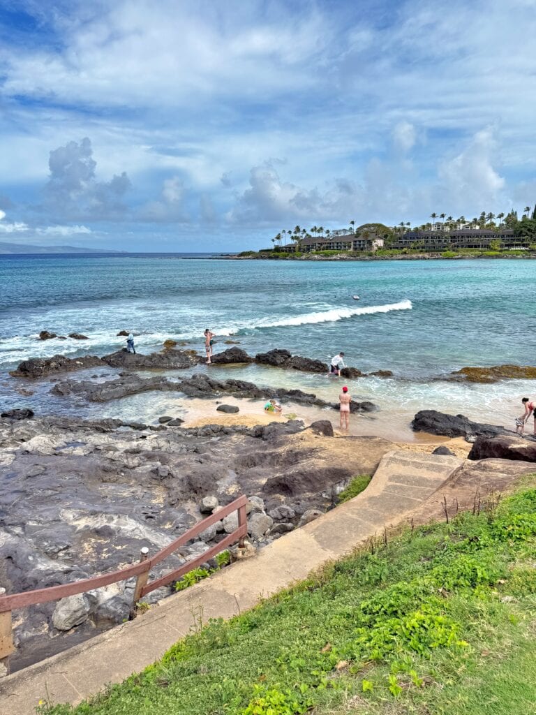 A rocky shoreline with several people by the water, waves breaking near the rocks, and buildings and palm trees in the background under a partly cloudy sky—perfect for Napili Bay snorkeling on Maui.