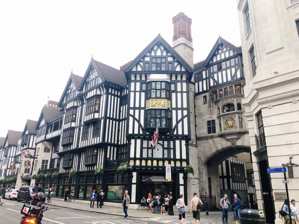 A large Tudor-style building with black and white timber framing, an archway, and people walking on the street in front.