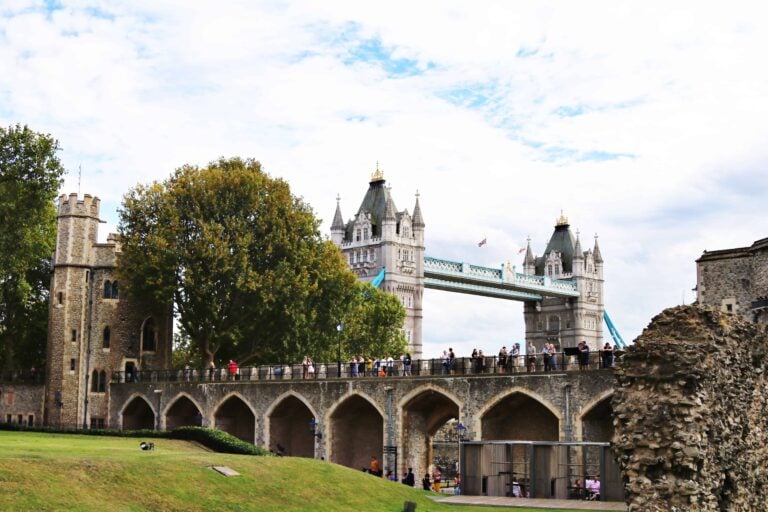 A group of people stands on a stone walkway with the Tower Bridge and historic buildings visible in the background under a partly cloudy sky.