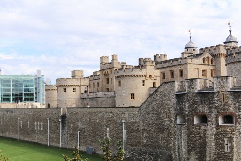 A large stone castle with towers and battlements stands behind a high stone wall; a modern glass building is visible in the background.