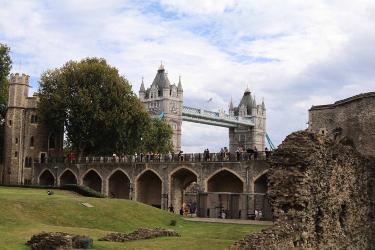 View of tourists walking along a stone walkway at the Tower of London, with Tower Bridge visible in the background under a partly cloudy sky.