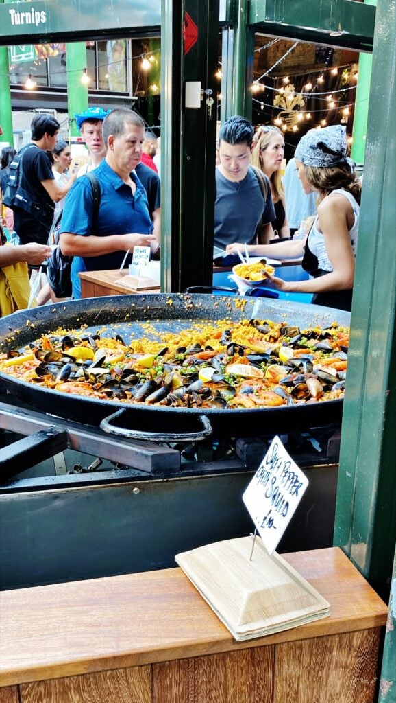 Large pan of seafood paella at a market stall, with people in the background serving themselves and waiting in line. A sign in front reads "Seafood Paella." It’s a delicious highlight among many London things to do for free.