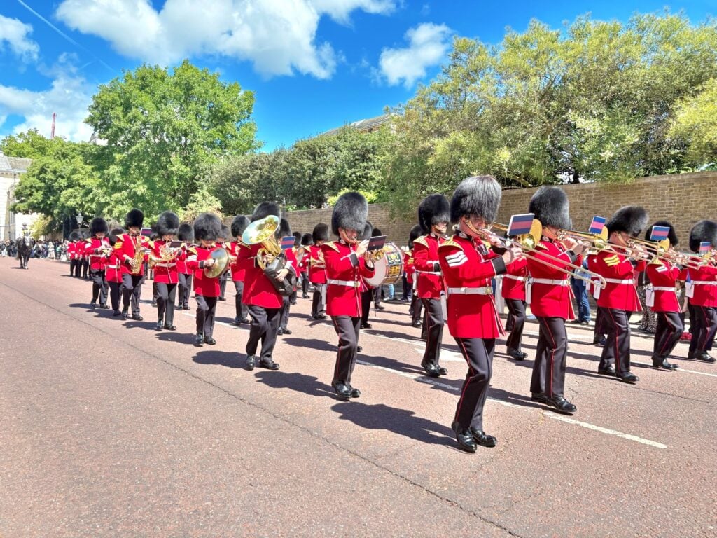 A marching band in red uniforms and bearskin hats performs on a sunny street, playing brass instruments—one of the classic London things to do for free—with trees and a stone wall in the background.