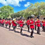 A marching band in red uniforms and bearskin hats performs on a sunny street, playing brass instruments—one of the classic London things to do for free—with trees and a stone wall in the background.