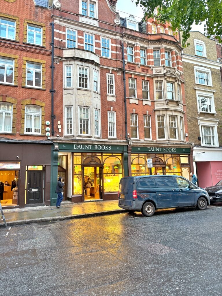 A street view of Daunt Books, a bookstore with lit windows on the ground floor of a red brick building, with a parked van and a person walking by—one of the classic London things to do for free.