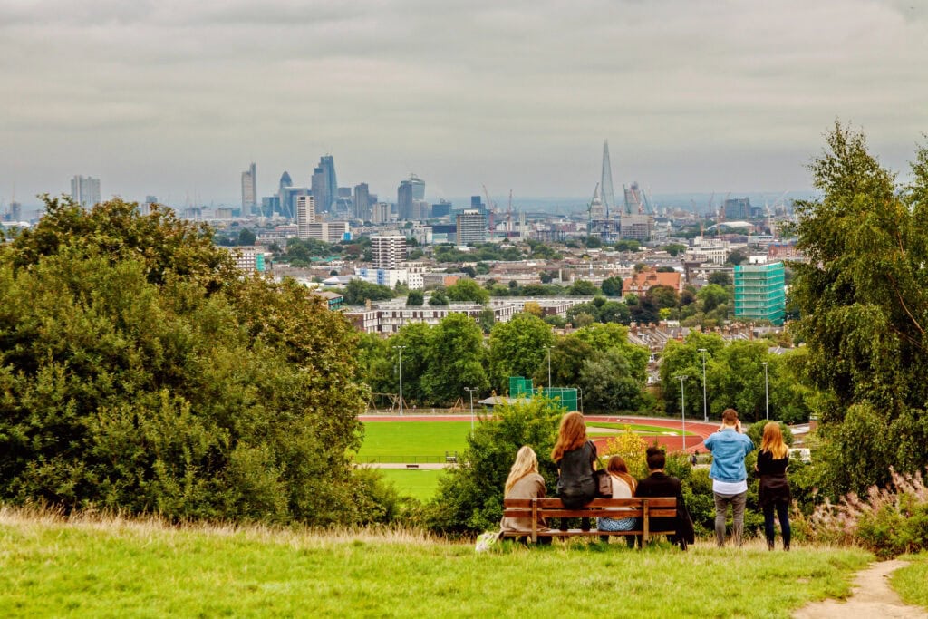 A group of people sit and stand by a bench on a grassy hill overlooking the London skyline—a perfect example of London things to do for free, enjoying stunning city views without spending a penny.