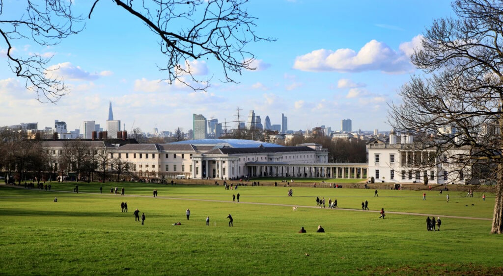 People walk and relax on a large grassy field with historic white buildings and the London skyline, including The Shard, visible in the background—a perfect spot among London things to do for free under a partly cloudy sky.