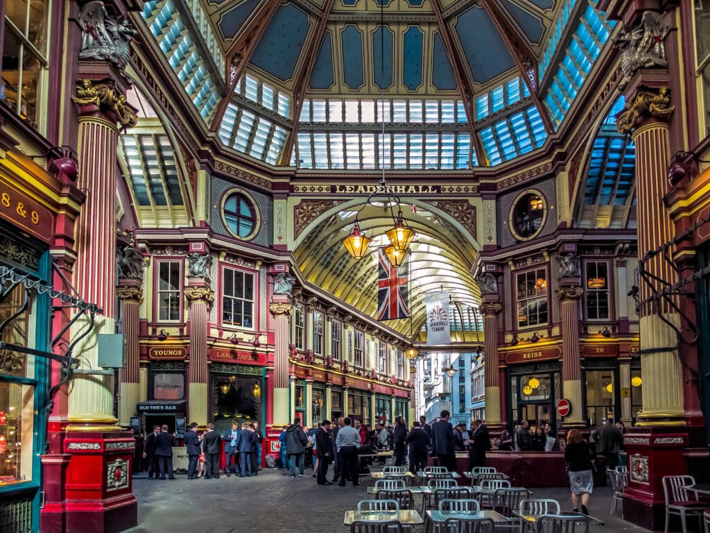 Indoor market hall with ornate architecture, arched glass roof, British flag, and people gathered among shops and cafes; empty chairs in the foreground—one of the top London things to do for free.