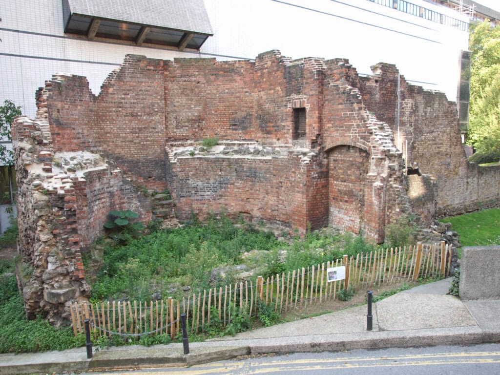 Remains of an old brick and stone wall with overgrown vegetation inside, bordered by a low wooden fence, next to a street and modern buildings—one of many London things to do for free.