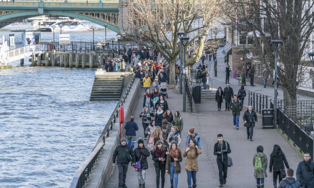 People walk along a riverside path near a bridge on a clear day, with some trees lining the walkway and water visible on the left—one of many London things to do for free.