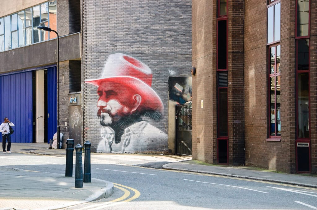 A mural of a man in a white hat with red accents is painted on a brick wall near a street corner—one of the unique London things to do for free in this vibrant urban area.