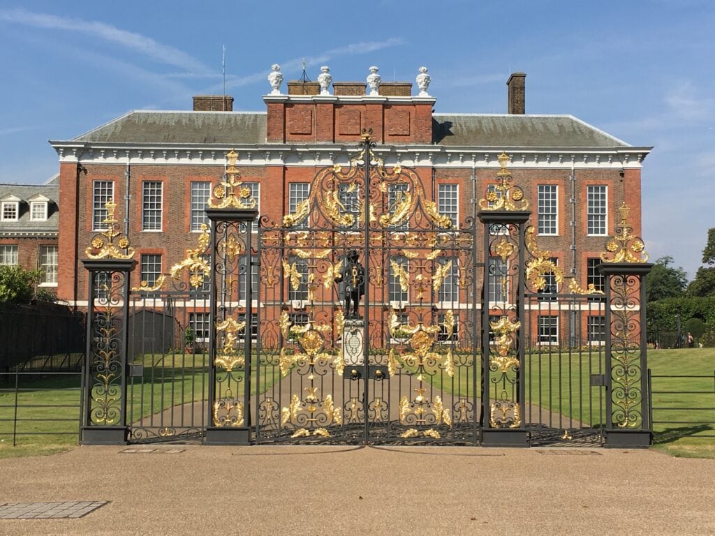 Ornate gold and black iron gates in front of a large red-brick historic building under a clear blue sky.