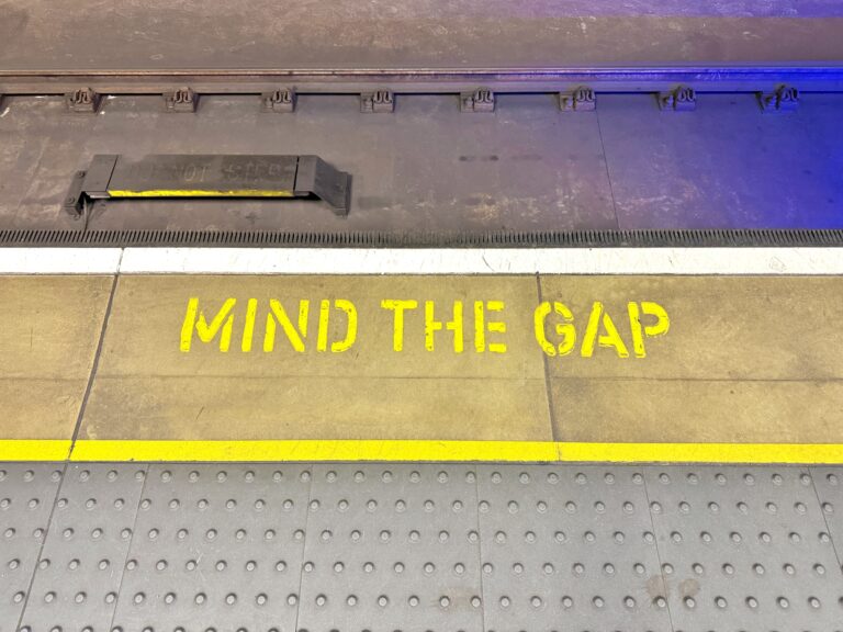 Yellow "MIND THE GAP" text painted on a subway platform edge, with tactile paving in front and train tracks behind.