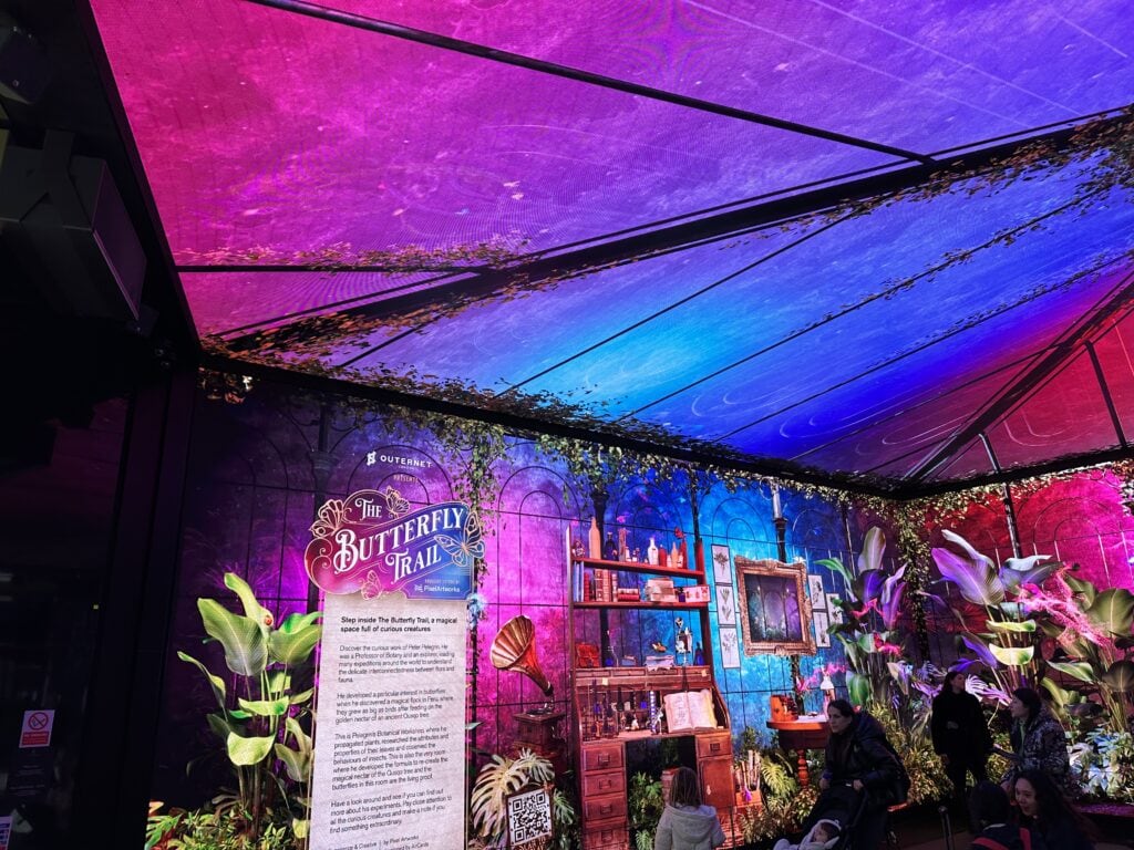 Colorful indoor display titled “The Butterfly Trail” with tropical plants, vintage decor, and an information panel under a purple and blue illuminated canopy—one of the unique London things to do for free.