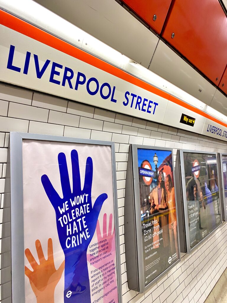 Liverpool Street Underground station platform features posters—including guidance on not tolerating hate crime—and clear signage for directions, making it easy for travelers learning how to use London tube services.