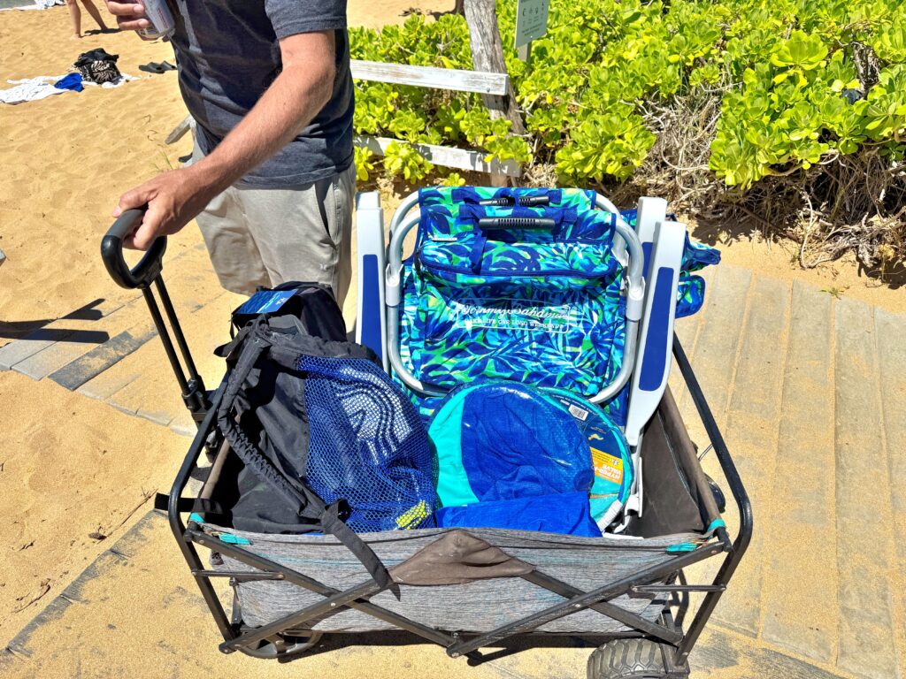A person pulls a wagon filled with beach gear—including folding chairs, a blue bag, and a towel—on a sandy path next to green shrubs, capturing the perfect scene for any Maui Travel Guide for First Timers.