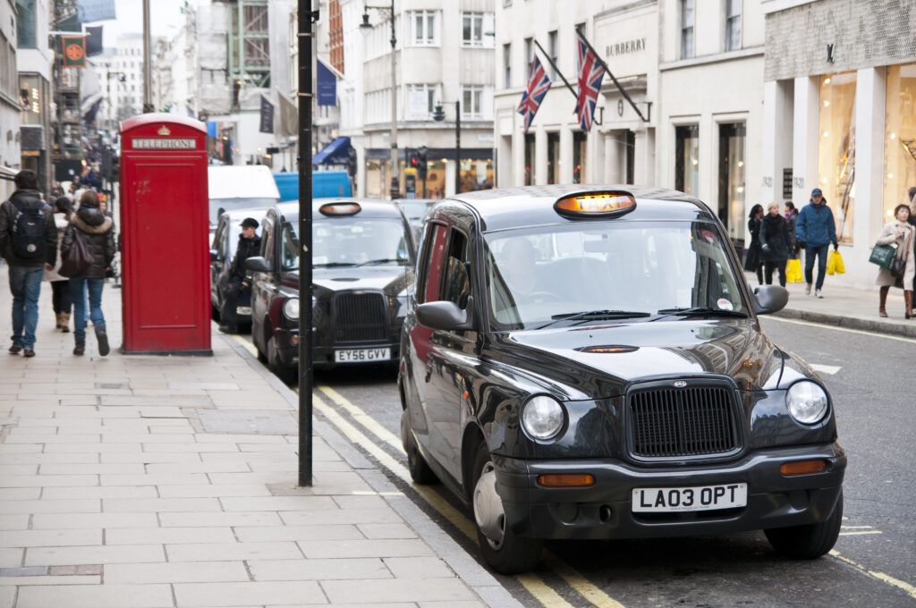 Black London taxis are parked by the curb on a city street near a red phone booth, with pedestrians walking and shops lining the road—perfect for exploring before you learn how to use the London tube.