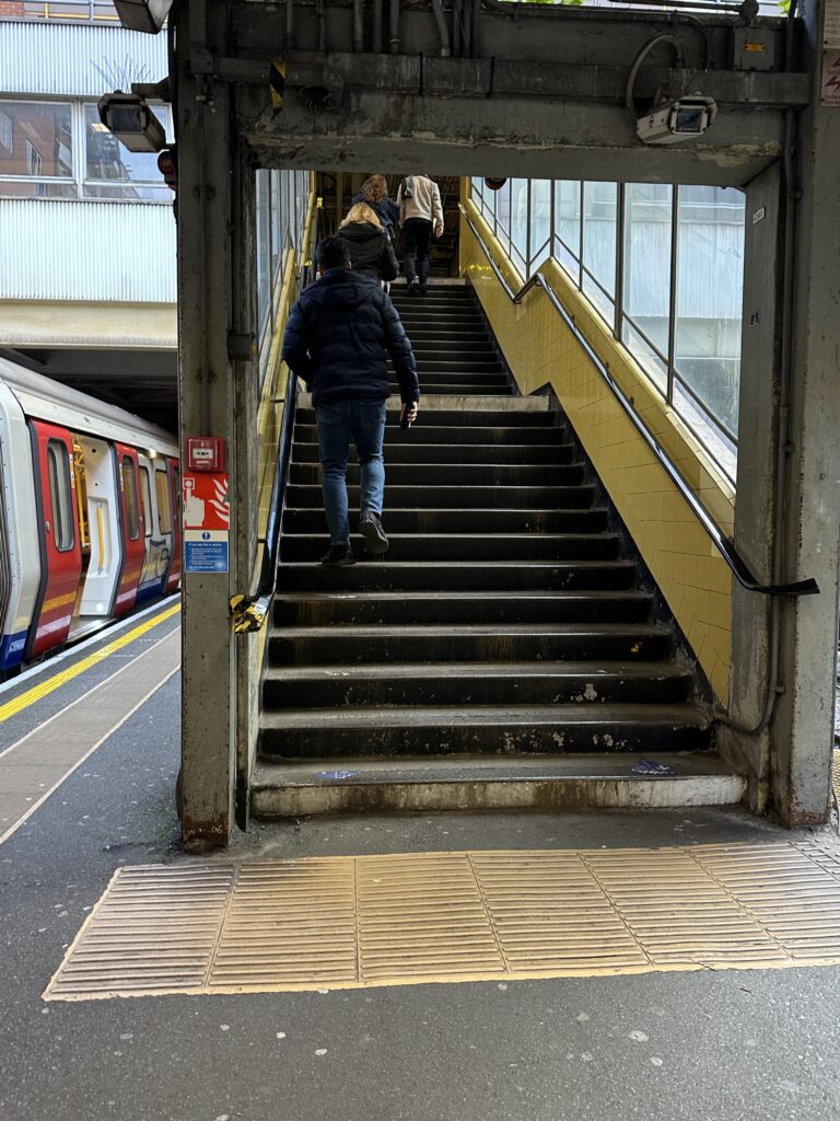 Two people walk up a staircase at a train station, with a train stopped on the platform to the left and yellow tactile paving on the ground—similar to scenes you might find when learning how to use London Tube stations.