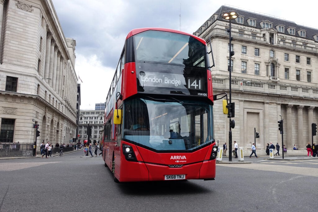 A red double-decker London bus on route 141 to London Bridge drives through a city intersection, with pedestrians and historic buildings nearby—a classic scene for visitors learning how to use the London Tube and buses.