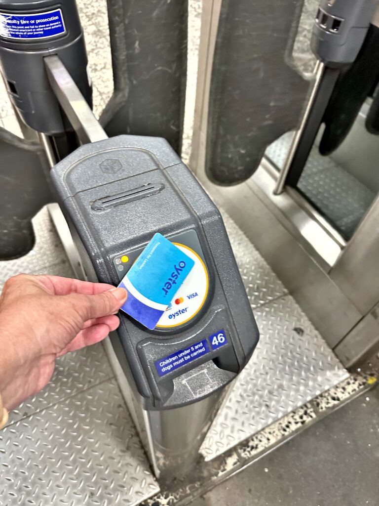 A person holds an Oyster card against a card reader at a subway turnstile, demonstrating how to use London Tube by tapping in for entry.
